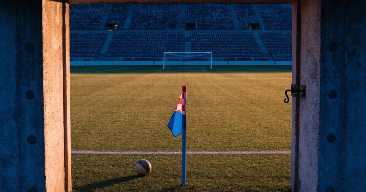 Blick durch ein Stadiontor auf einen grünen Rasen im Abendlicht, ein Ball ruht an der Eckfahne, die Tribünen sind leer.